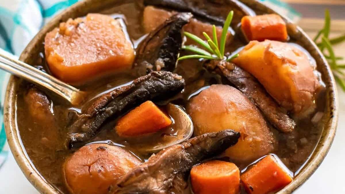 A bowl of hearty stew filled with chunks of potatoes, carrots, and mushrooms, garnished with a sprig of rosemary. The stew appears rich and flavorful, with a deep brown broth. A spoon rests in the bowl.