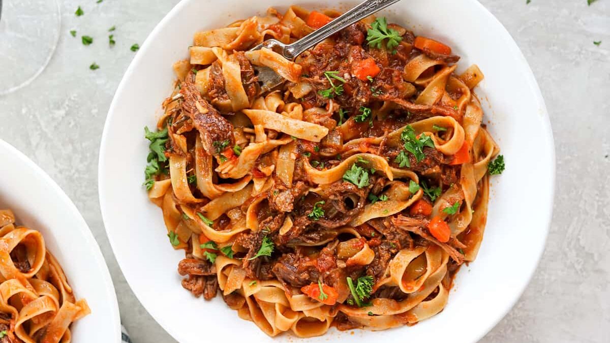 A bowl of wide pasta noodles topped with a rich, red sauce containing shredded meat, diced tomatoes, and fresh parsley. A fork rests in the bowl, and the dish is garnished with sprigs of parsley. Another dish is partially visible on the side.