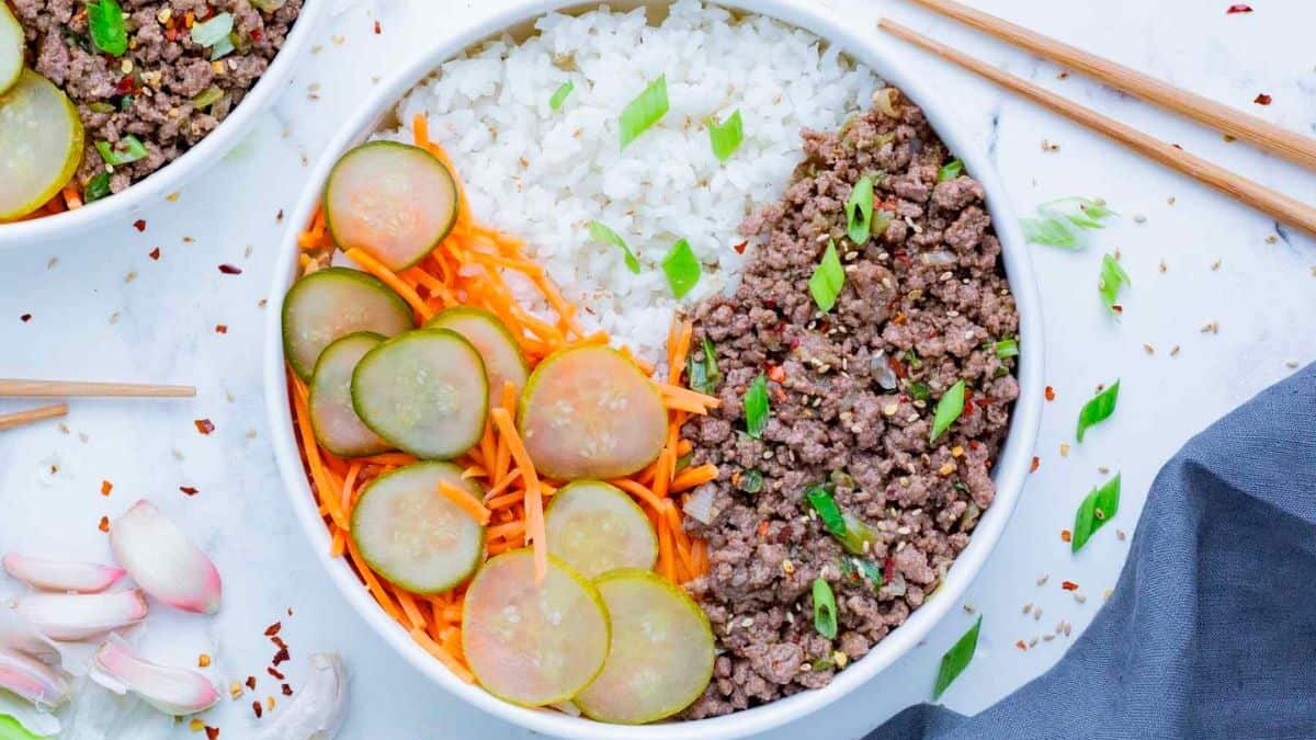 A bowl filled with ground beef, white rice, sliced pickles, and shredded carrots, garnished with sliced green onions. The bowl is surrounded by chopsticks, garlic cloves, and red pepper flakes on a white surface.