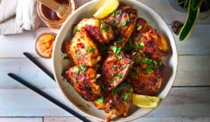 A bowl of glazed, grilled chicken pieces garnished with chopped herbs and served with lemon wedges. Nearby are chopsticks, a jar of sauce, and a small jar of pickles on a light wooden surface.
