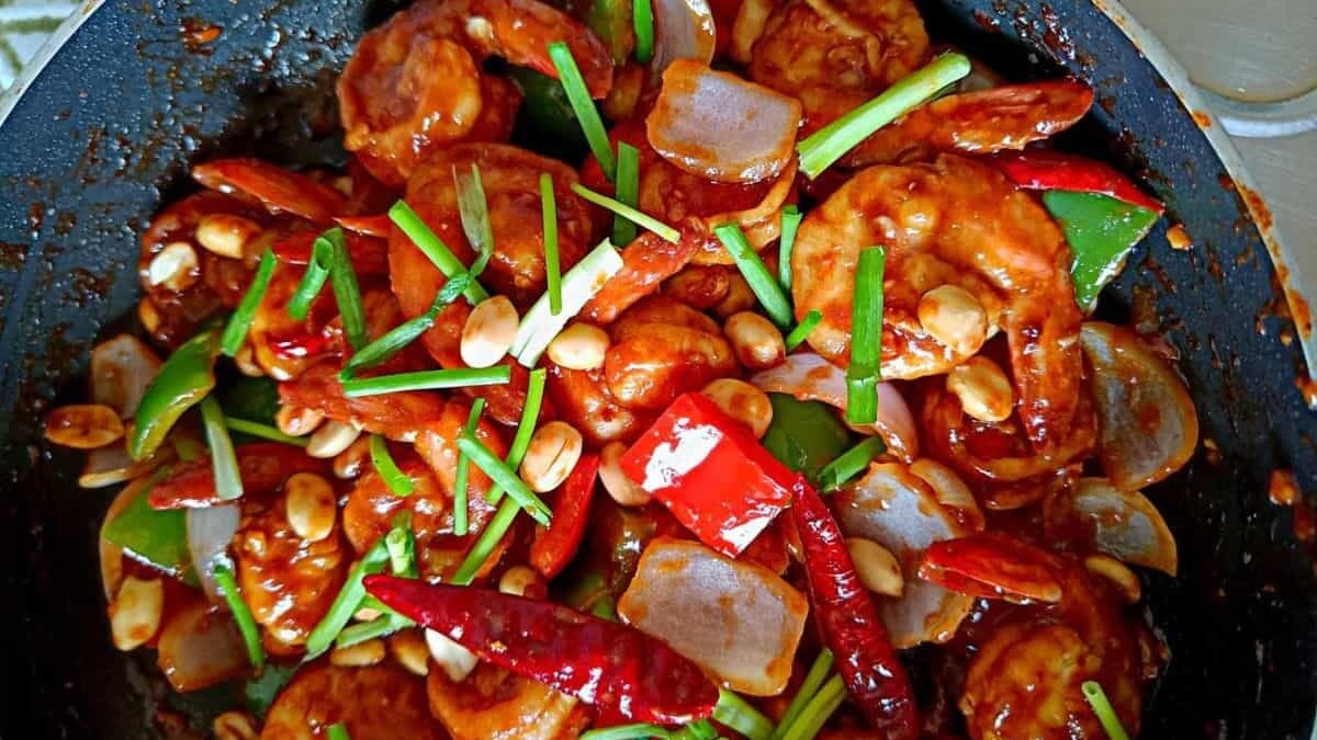 A stir-fry dish in a pan features prawns, red and green bell peppers, onions, peanuts, and dried red chilies, all coated in a glossy sauce. Garnished with green onion slices.