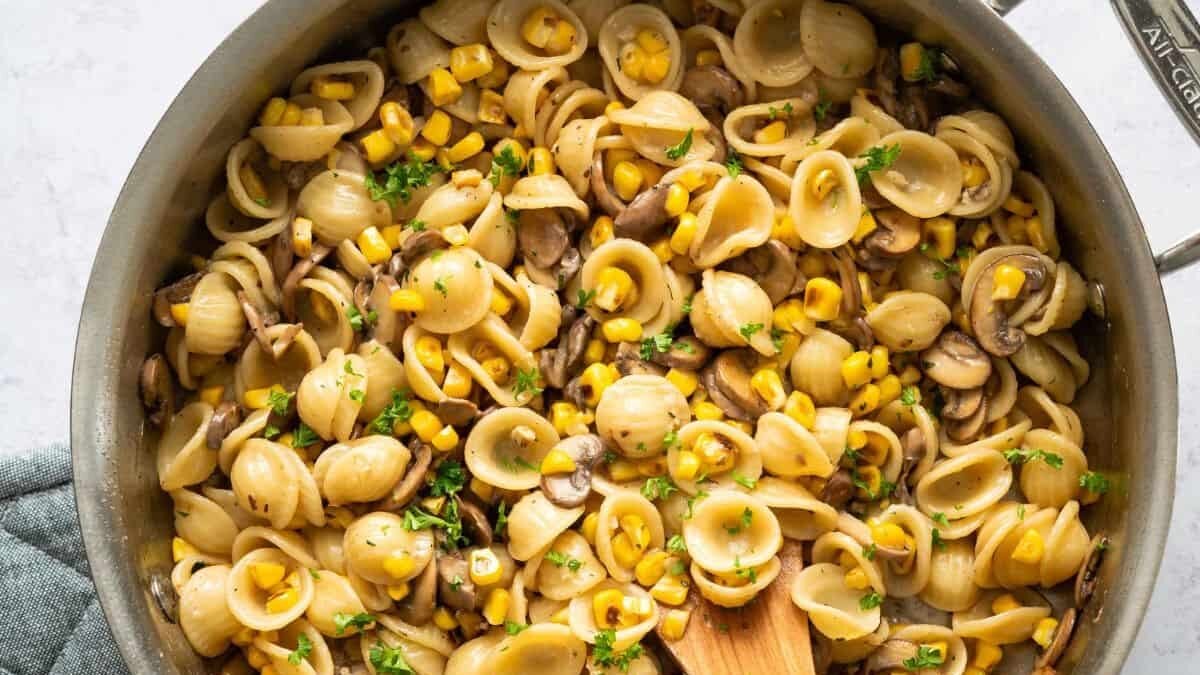 A skillet filled with orecchiette pasta, corn, and sliced mushrooms, garnished with fresh parsley. A wooden spoon rests inside, ready to serve.