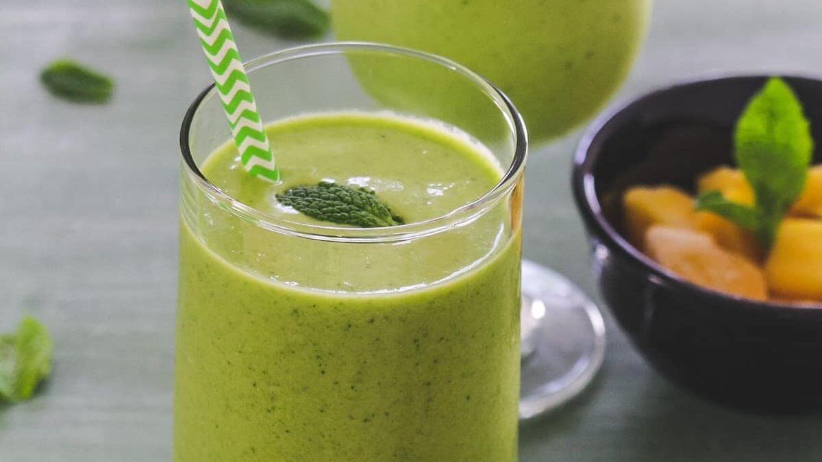 A glass of green smoothie with a striped straw and mint garnish sits on a table. A black bowl containing sliced fruit is in the background. The smoothie is smooth and vibrant.