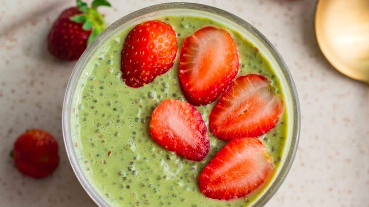 A bowl of green smoothie topped with sliced strawberries and chia seeds. A few whole strawberries are placed in the background on a light marble surface.