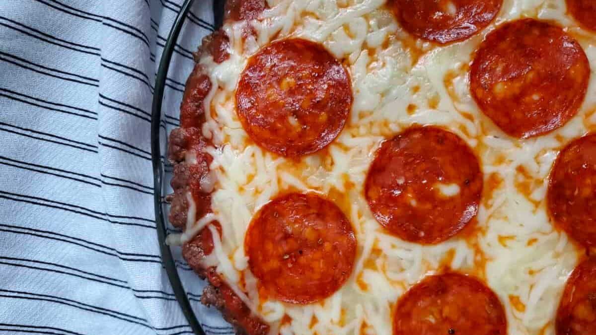 Close-up of a pepperoni pizza with melted cheese and pepperoni slices on a dark pan. A striped cloth is partially visible beneath the pan.