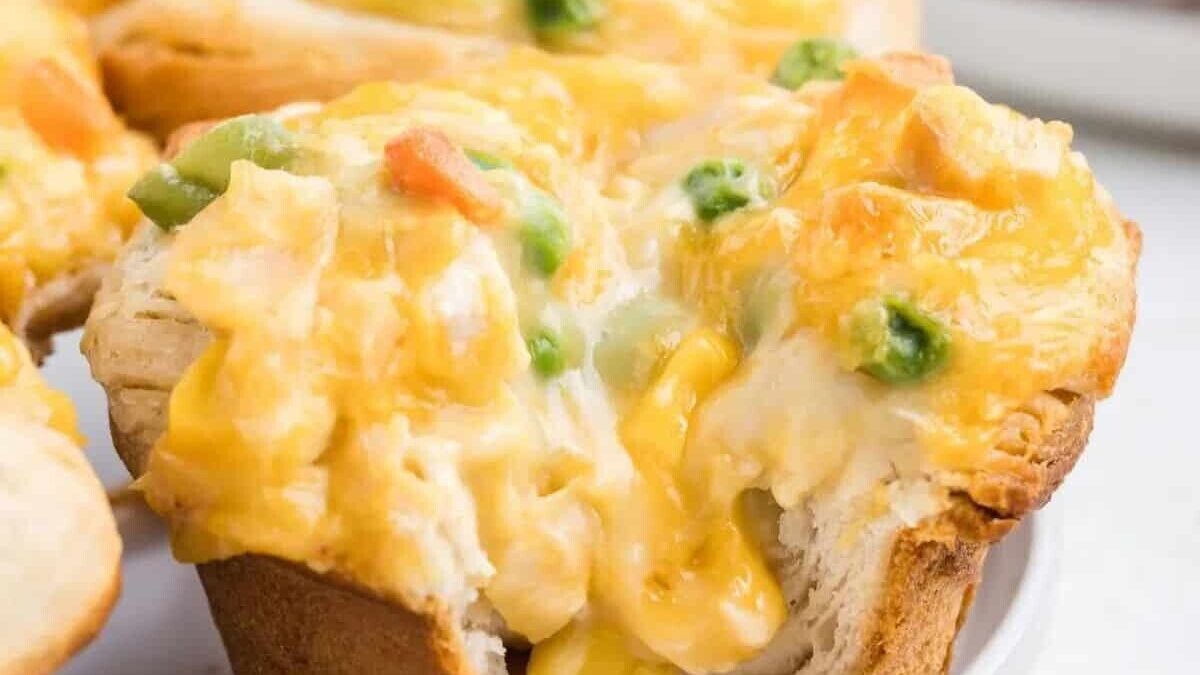 A savory muffin filled with creamy chicken, melted cheese, and pieces of colorful vegetables like peas and carrots, showing a gooey interior, placed on a white plate.