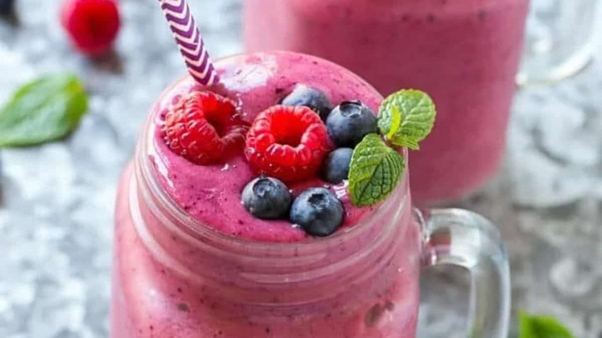 A mason jar filled with a vibrant pink berry smoothie, topped with fresh raspberries, blueberries, and a sprig of mint, with a purple-striped straw, on a gray surface.