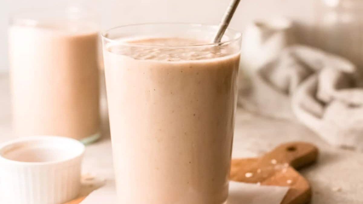A creamy smoothie in a clear glass with a metal straw, placed on a wooden board. In the background, there is a blurred glass filled with the same drink and a small white dish. A light-colored cloth is partially visible.