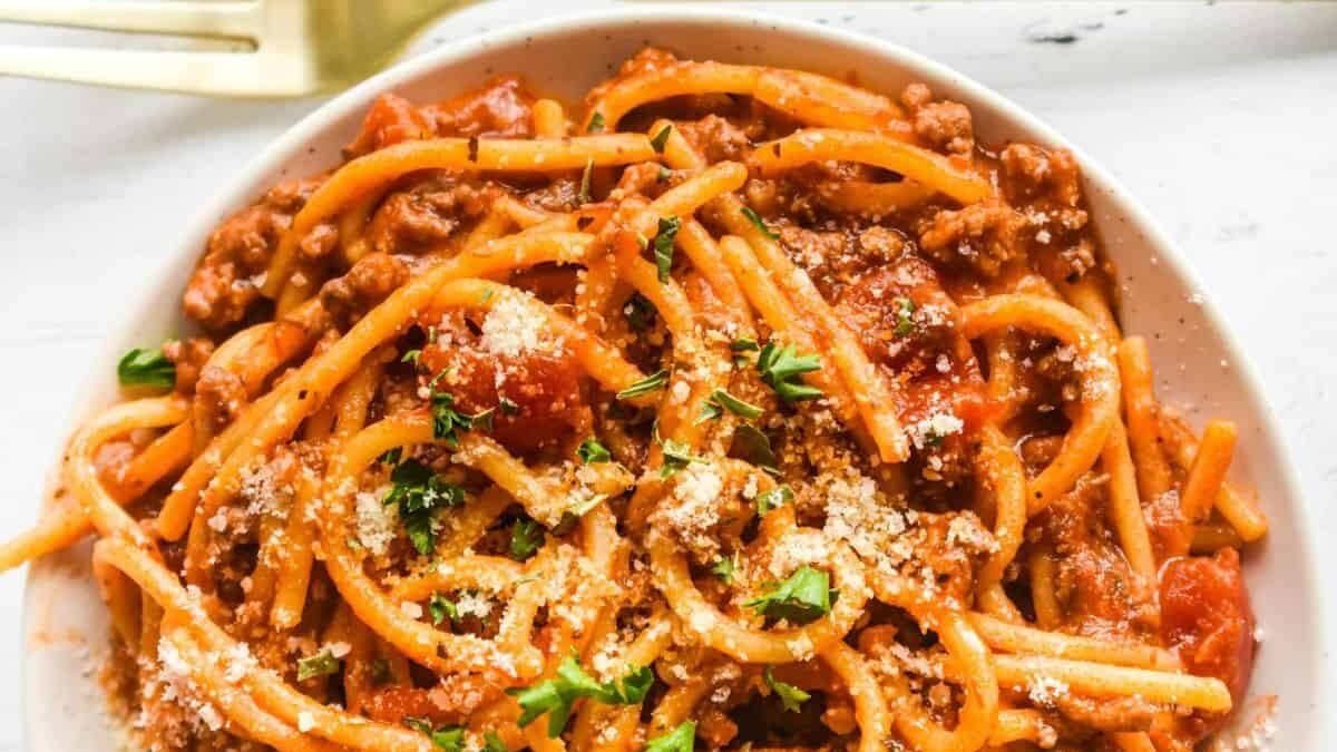 A bowl of spaghetti topped with ground meat tomato sauce, garnished with grated cheese and parsley. A fork is visible on the side, resting on a white surface.