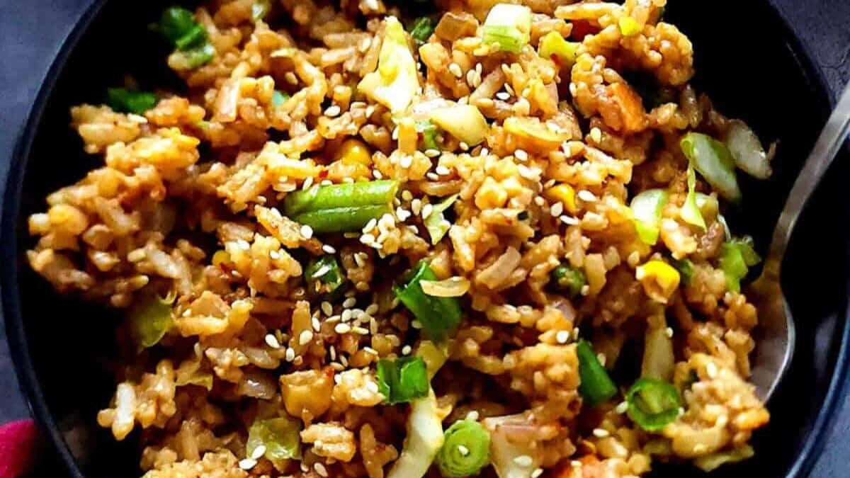 A bowl of fried rice with vegetables, topped with sesame seeds. The dish includes green onions, corn, and cabbage, served with a spoon.