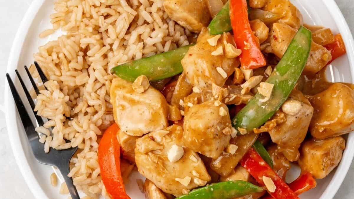 A plate of brown rice and stir-fried chicken with red bell peppers, snow peas, and peanuts in a sauce. A fork is placed beside the rice.