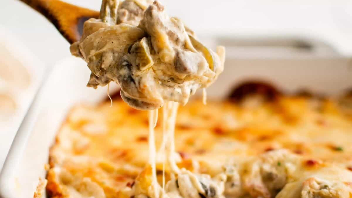 A wooden spoon lifts a cheesy, creamy pasta bake from a white baking dish. The dish is topped with melted cheese and has visible pieces of mushrooms and other ingredients, creating a savory, gooey texture.