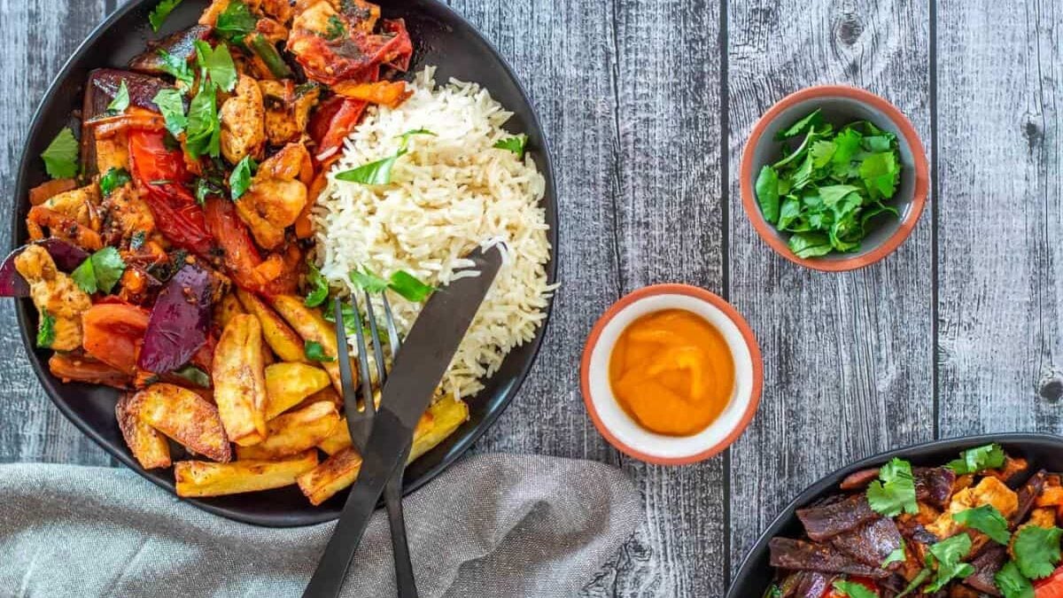 A colorful meal is spread out on a wooden table. Each black plate contains stir-fried vegetables, golden-fried potatoes, and a portion of white rice. Nearby are bowls of fresh herbs and a yellow sauce. Two forks and a knife rest on a cloth napkin beside a plate.