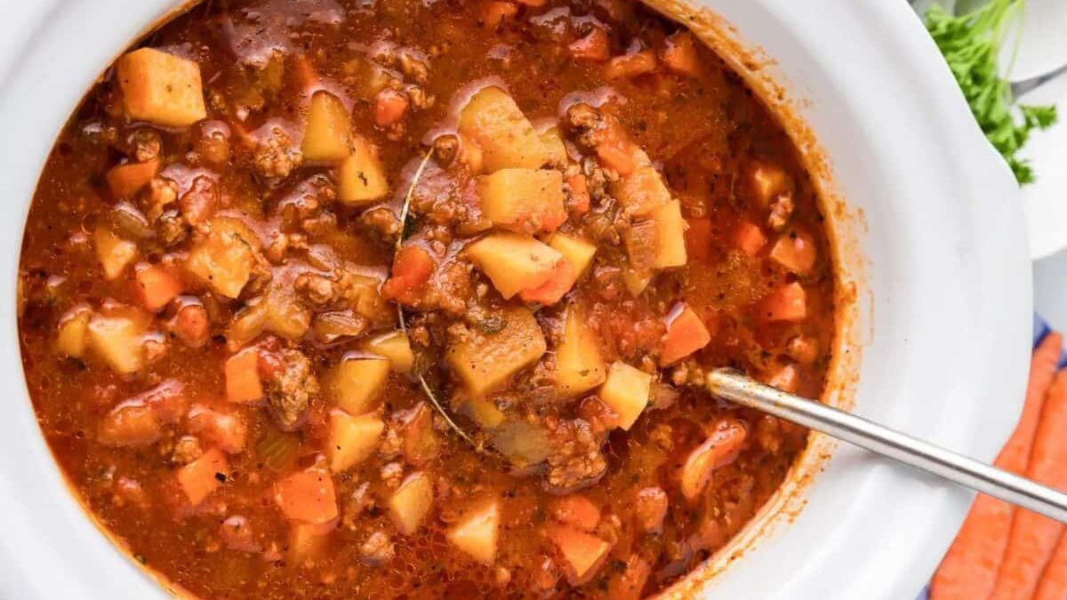 A close-up of a hearty beef stew in a white pot, featuring chunks of beef, potatoes, and carrots in a rich, brown broth. A metal ladle rests in the stew, ready to serve. Fresh carrots and parsley garnish the background.