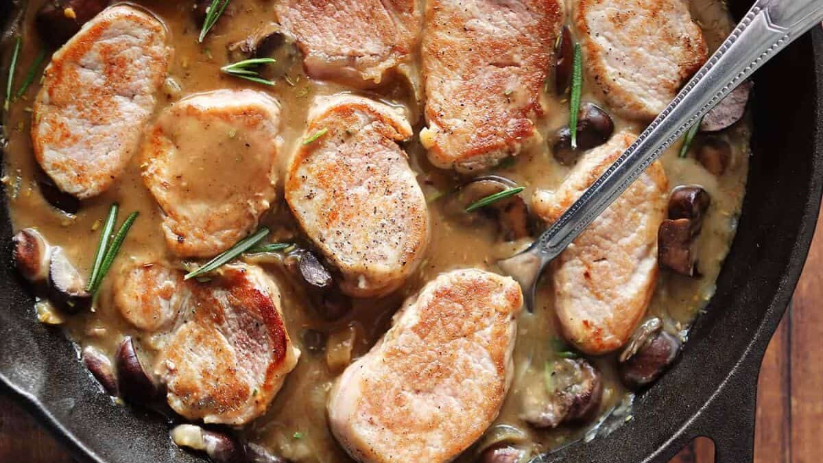 Pan-seared pork chops in a creamy sauce with mushrooms and rosemary sprigs in a skillet, accompanied by a silver serving spoon.