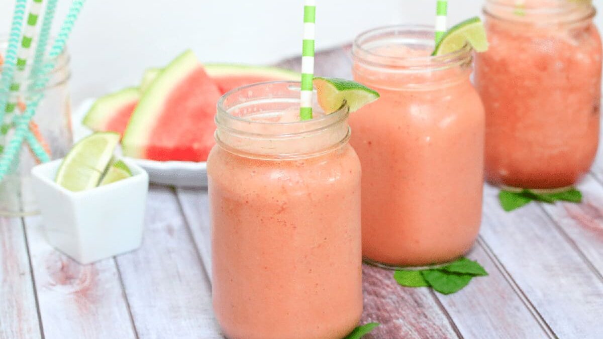 Three jars of watermelon smoothies with lime slices and green-striped straws are on a wooden table. In the background, there are watermelon slices and extra lime wedges.