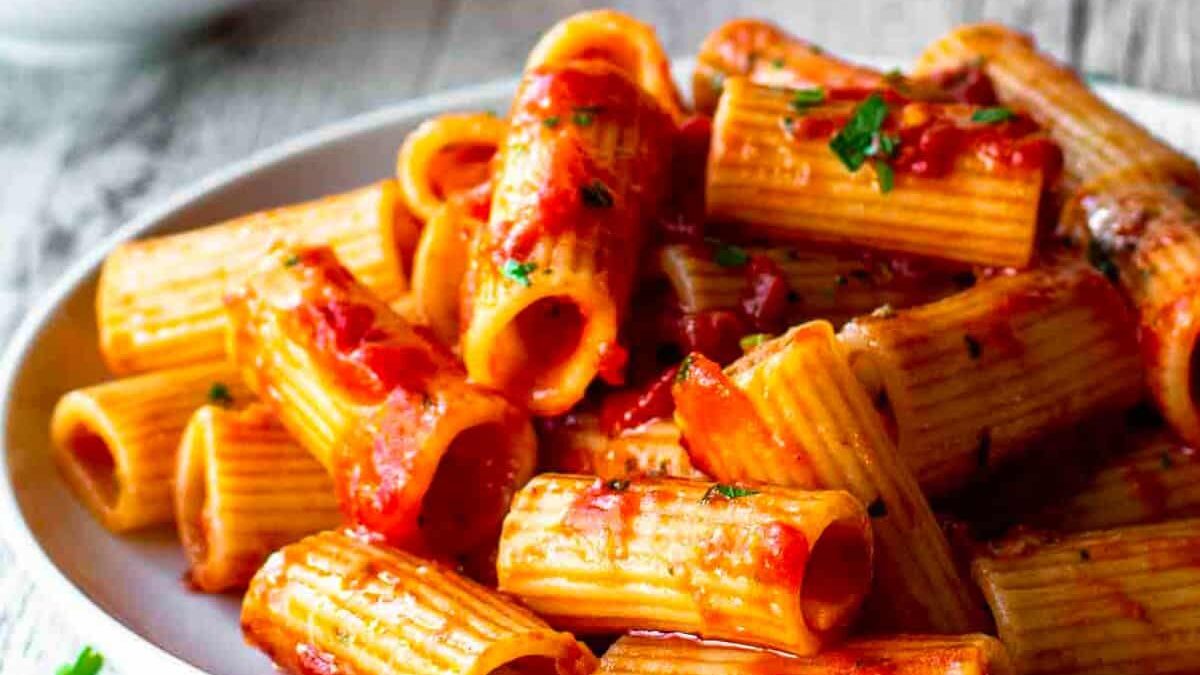 A plate of rigatoni pasta topped with a rich, red tomato sauce. The pasta is garnished with chopped herbs, adding a touch of green. The dish is served on a white plate, set on a rustic wooden table.