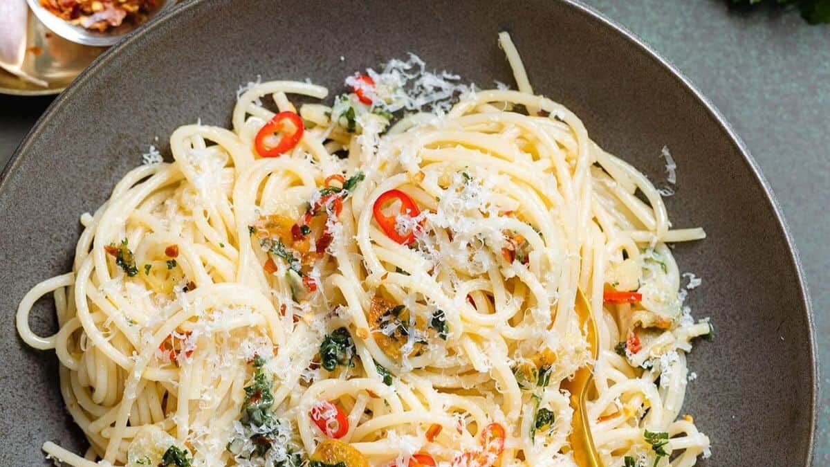 A bowl of spaghetti aglio e olio topped with grated cheese, sliced red chili peppers, and parsley. A fork is tucked into the pasta. A small dish of red pepper flakes is visible in the background.