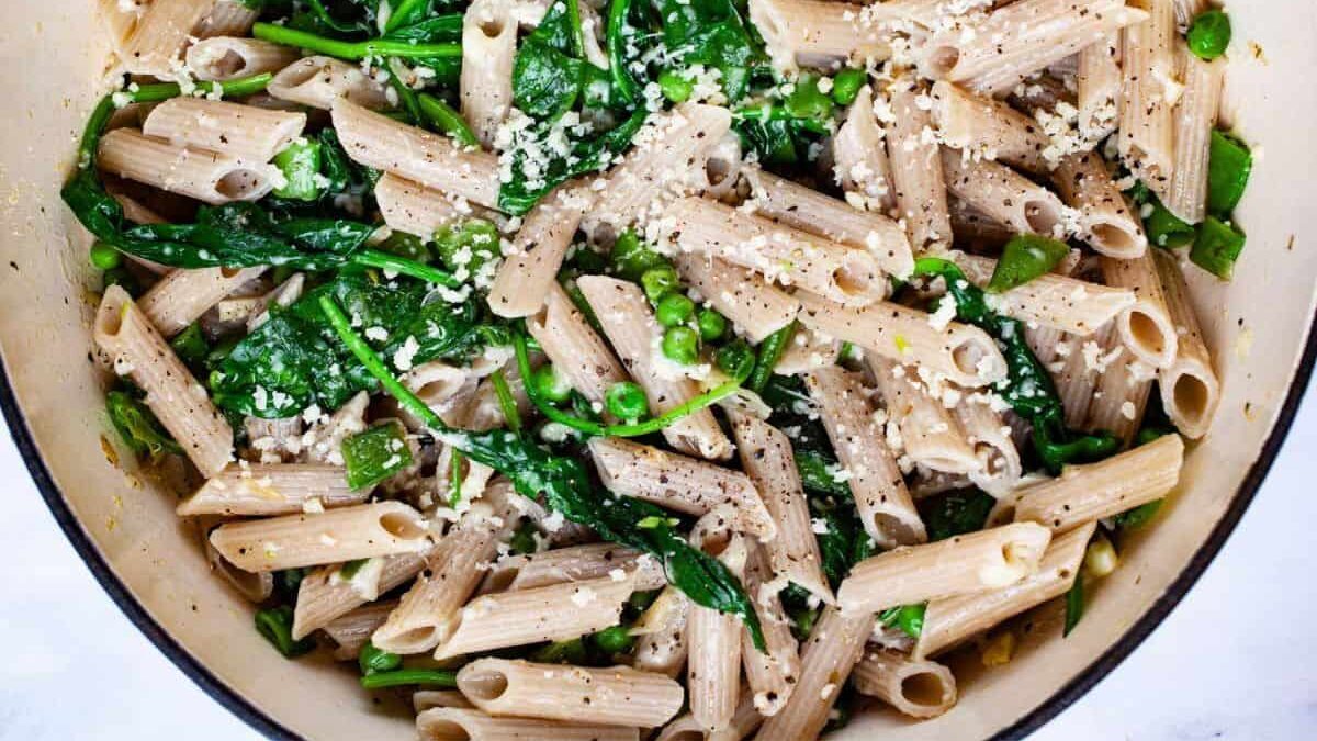 A bowl of whole wheat penne pasta mixed with peas, spinach, and green beans, topped with grated cheese and black pepper.