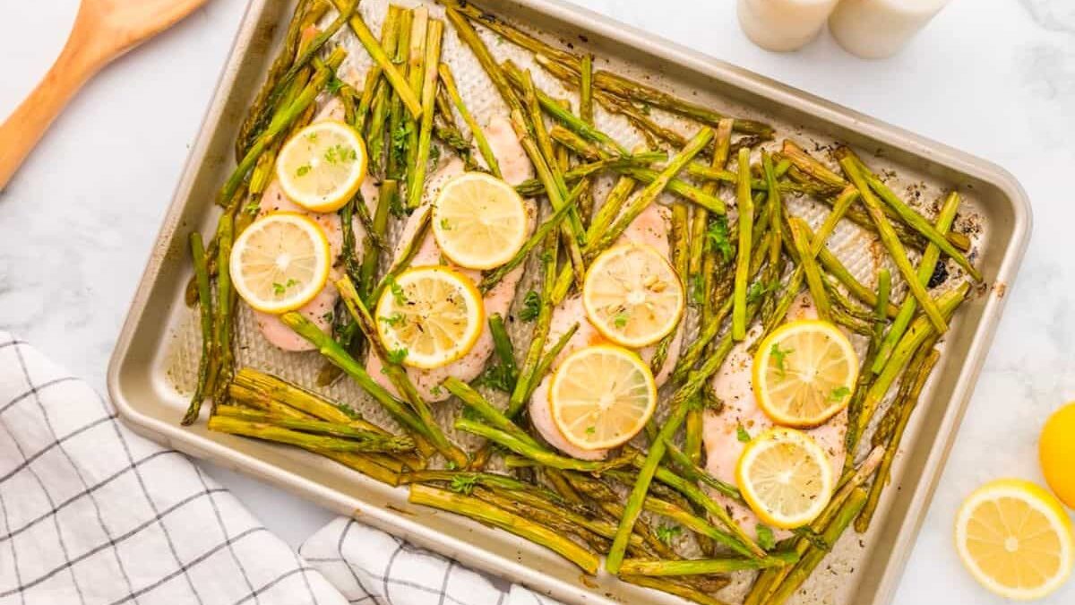 Baked salmon fillets on a tray, topped with lemon slices and garnished with parsley, surrounded by roasted asparagus. A wooden spoon and checkered cloth are beside the tray on a marble surface.
