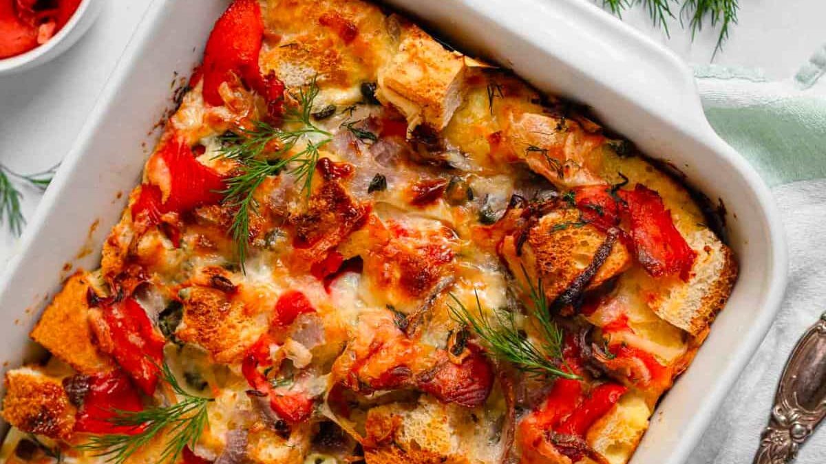 A close-up of a baked dish in a white casserole. It contains golden-brown bread cubes, red bell pepper pieces, melted cheese, and sprigs of dill. A small bowl of red sauce and dill fronds are visible in the background.