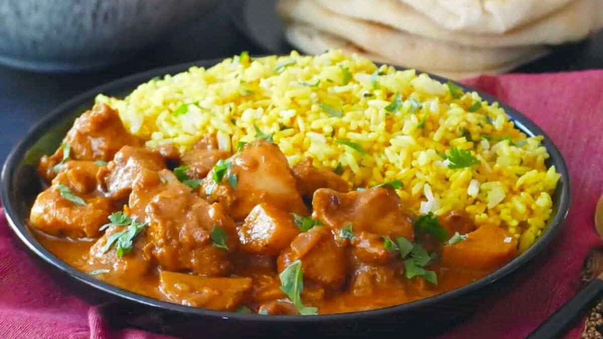 A plate of curry chicken and yellow rice garnished with fresh herbs. The dish is served on a black plate with slices of naan bread in the background, placed on a red cloth.