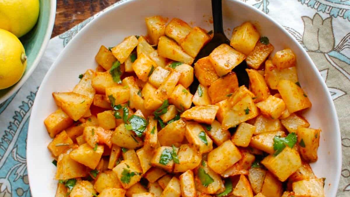A bowl of roasted potatoes seasoned with spices and garnished with chopped green herbs. A black spoon is resting in the bowl. The bowl is placed on a patterned tablecloth next to a plate of lemons.
