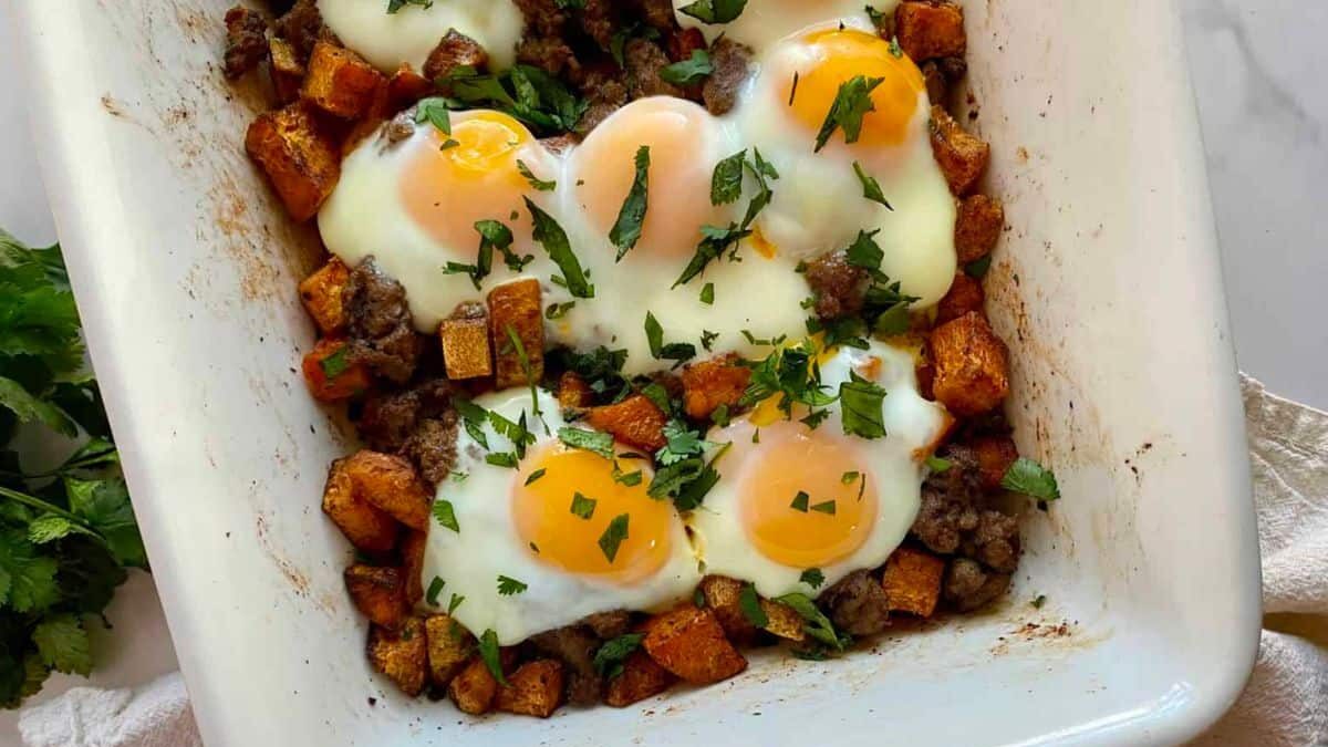A casserole dish filled with baked eggs on a bed of diced sweet potatoes and ground meat, garnished with chopped fresh herbs. It's placed on a white surface with a light-colored cloth and some green leaves nearby.