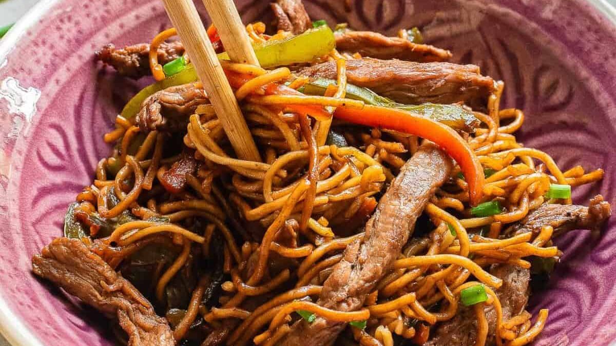 A bowl of stir-fried noodles with sliced beef, bell peppers, and green onions. Chopsticks rest on top, and the dish is served in a purple bowl. The noodles are coated in a savory sauce.