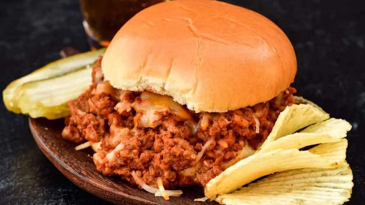 A sloppy joe sandwich with seasoned ground meat and melted cheese on a bun sits on a wooden plate. It's accompanied by wavy potato chips and pickle slices. A glass of a dark-colored beverage is in the background.