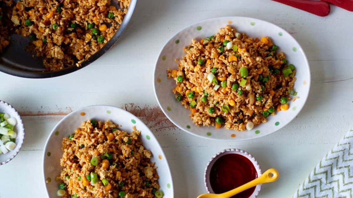 Three plates of fried rice mixed with peas, corn, and green onions are on a table. A bowl of red sauce with a yellow spoon is next to the plates. Two forks and a knife are placed beside the dishes. A skillet with more fried rice is partially visible.