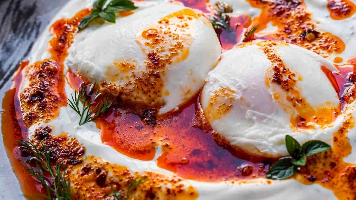 A creamy white yogurt dish topped with two poached eggs drizzled with spicy red oil, garnished with fresh herbs on a plate.