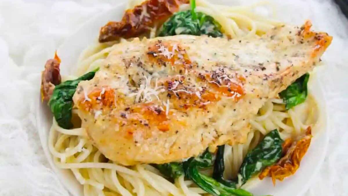 A seasoned, grilled chicken breast sits atop a bed of spaghetti, garnished with wilted spinach and sun-dried tomatoes. The dish is sprinkled with grated cheese, adding a touch of elegance to the creamy pasta.
