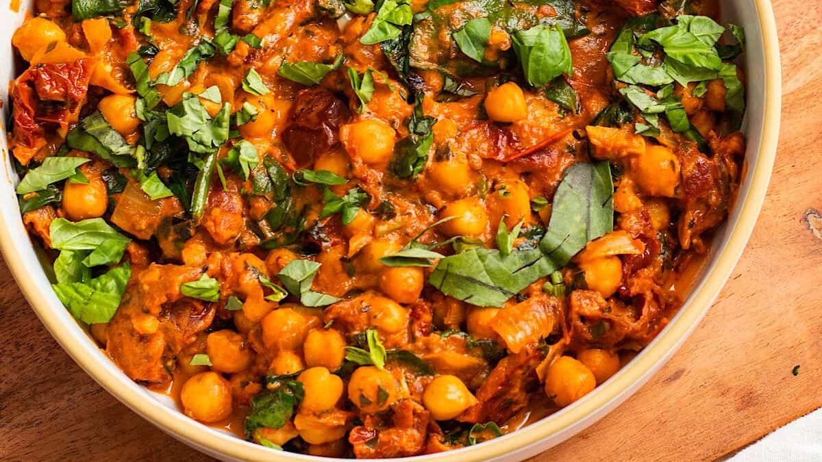 A close-up of a flavorful chickpea curry in a round dish. The curry is rich and creamy, filled with chickpeas, sun-dried tomatoes, and chopped spinach. The mixture is vibrant orange with green garnishes on top. It's placed on a wooden surface.
