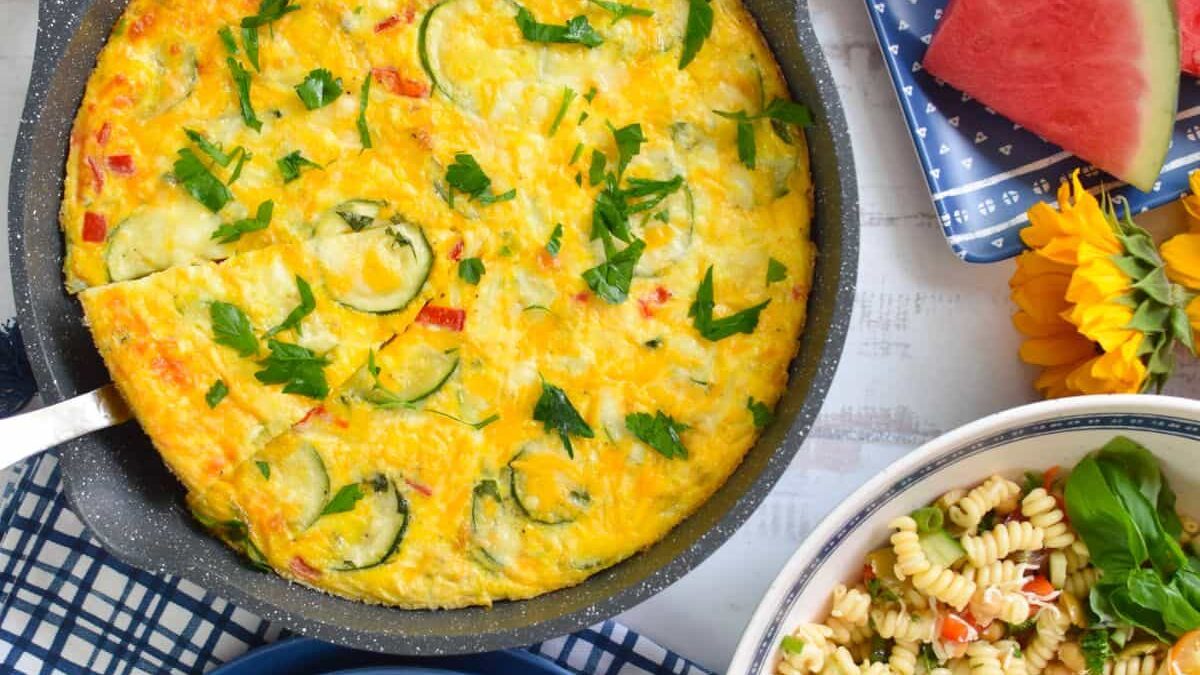 A colorful vegetable frittata in a black skillet garnished with herbs, accompanied by a plate of pasta salad with basil, slices of watermelon, and sunflowers on a white table.