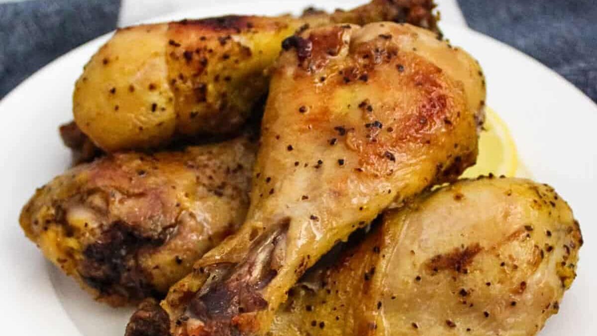 A plate of four golden-brown baked chicken drumsticks, seasoned with spices and black pepper. The chicken appears juicy and tender, and a white napkin is visible underneath part of the plate.