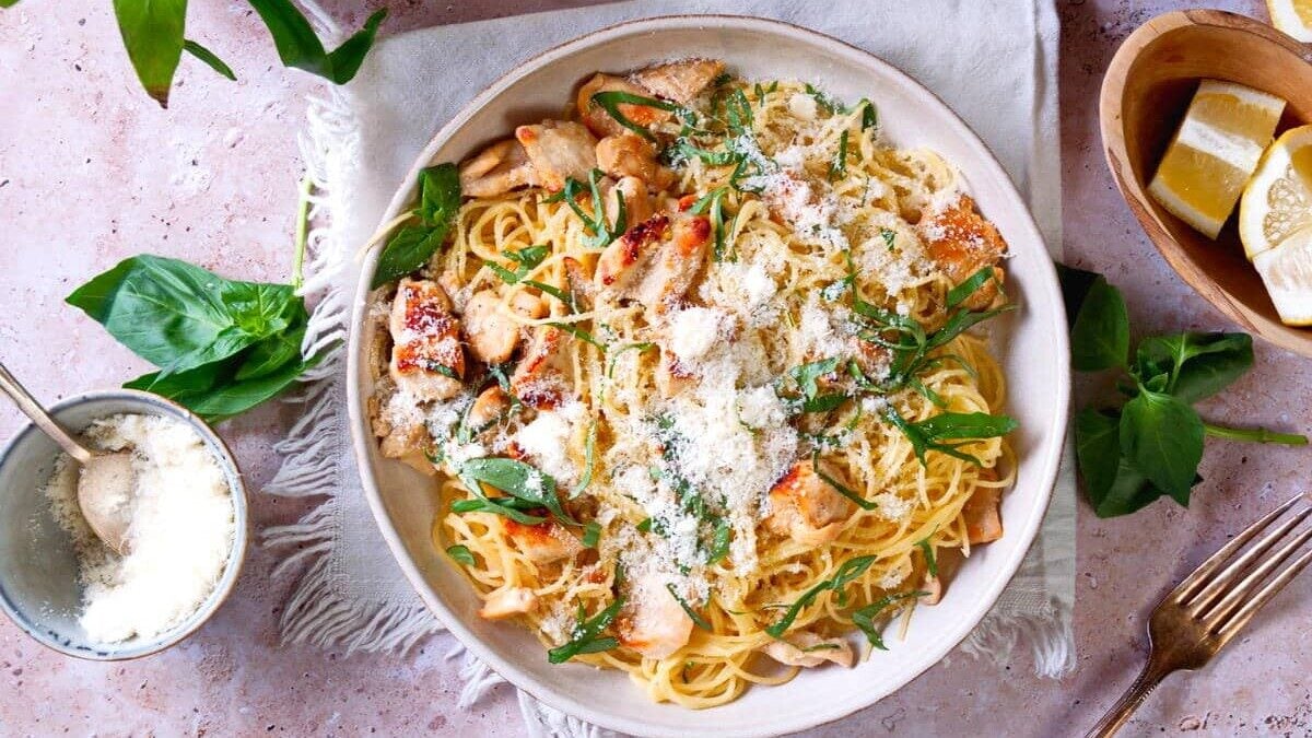 A bowl of pasta with grilled chicken, garnished with grated cheese and fresh herbs, placed on a white napkin. Surrounding the bowl are basil leaves, lemon wedges in a wooden bowl, a spoon with grated cheese, and a glass jar. A fork and knife sit beside the bowl.