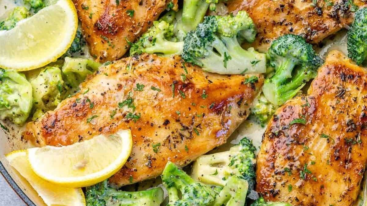 Grilled chicken breasts seasoned with herbs served with broccoli florets and lemon wedges, garnished with parsley. The dish is displayed in a pan, showcasing the golden brown chicken and vibrant green vegetables.