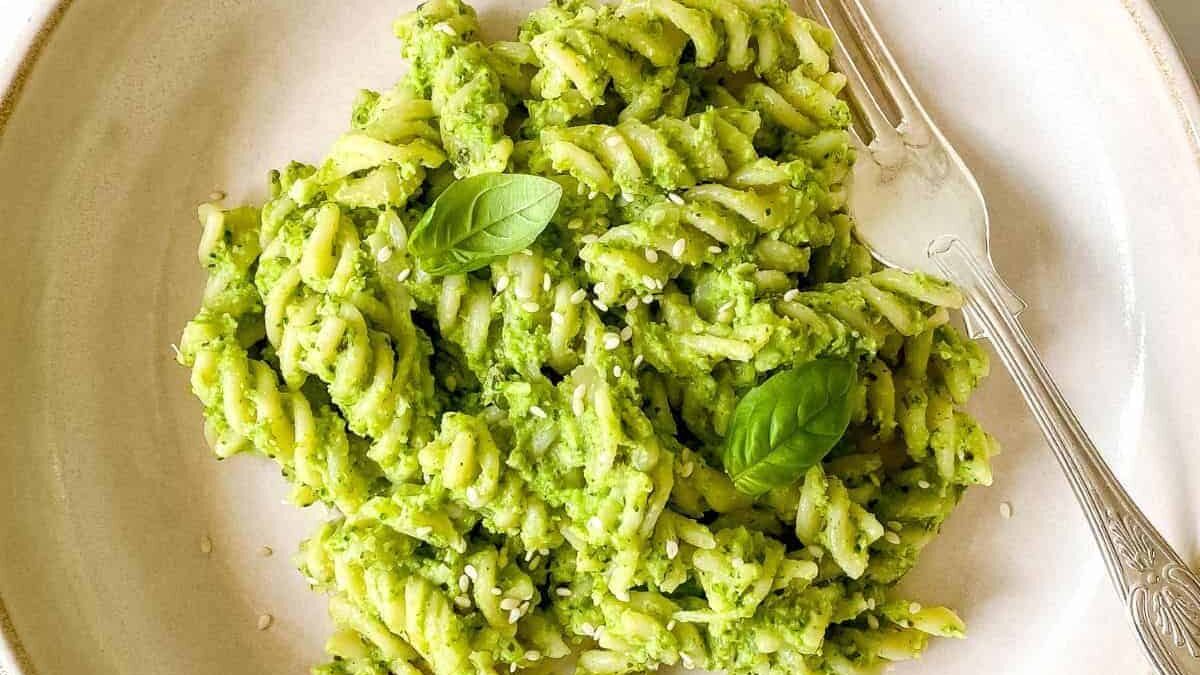 A plate of fusilli pasta covered in green pesto sauce, garnished with fresh basil leaves. A fork rests on the edge of the plate on a light-colored background.