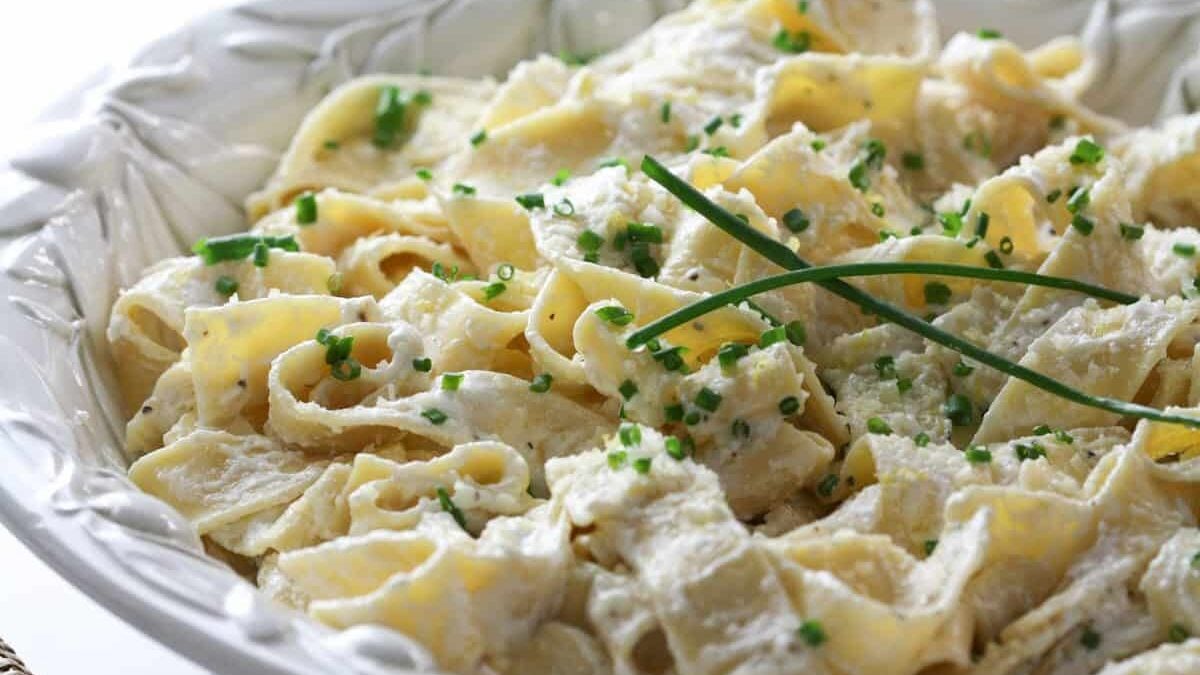 A close-up of a plate of fettuccine Alfredo garnished with chopped chives and two whole chive stems, served in a decorative white bowl.