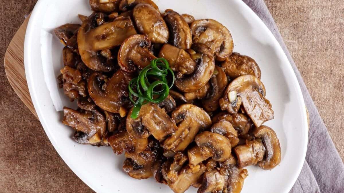 A white plate filled with sautéed mushrooms garnished with sliced green onions. The plate is set on a textured brown surface with a gray cloth partially visible.