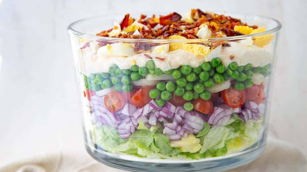 A glass bowl filled with layered salad. From bottom to top: shredded lettuce, chopped red onion, cherry tomato halves, green peas, diced boiled eggs, crispy bacon bits, and a thick creamy dressing. The layers are neatly stacked and vibrant.