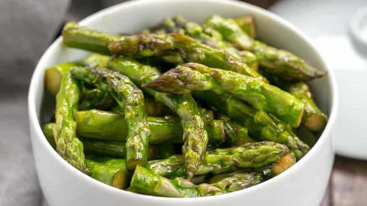 A white bowl filled with cooked asparagus spears, showcasing a glossy green color and slight char marks. The background is blurred, focusing attention on the asparagus.