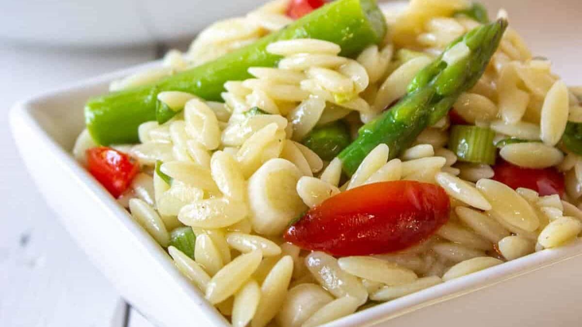 A square white dish filled with orzo pasta mixed with cherry tomatoes and asparagus. The pasta is lightly seasoned, and some pieces of chopped green onions are visible, enhancing the colorful presentation.