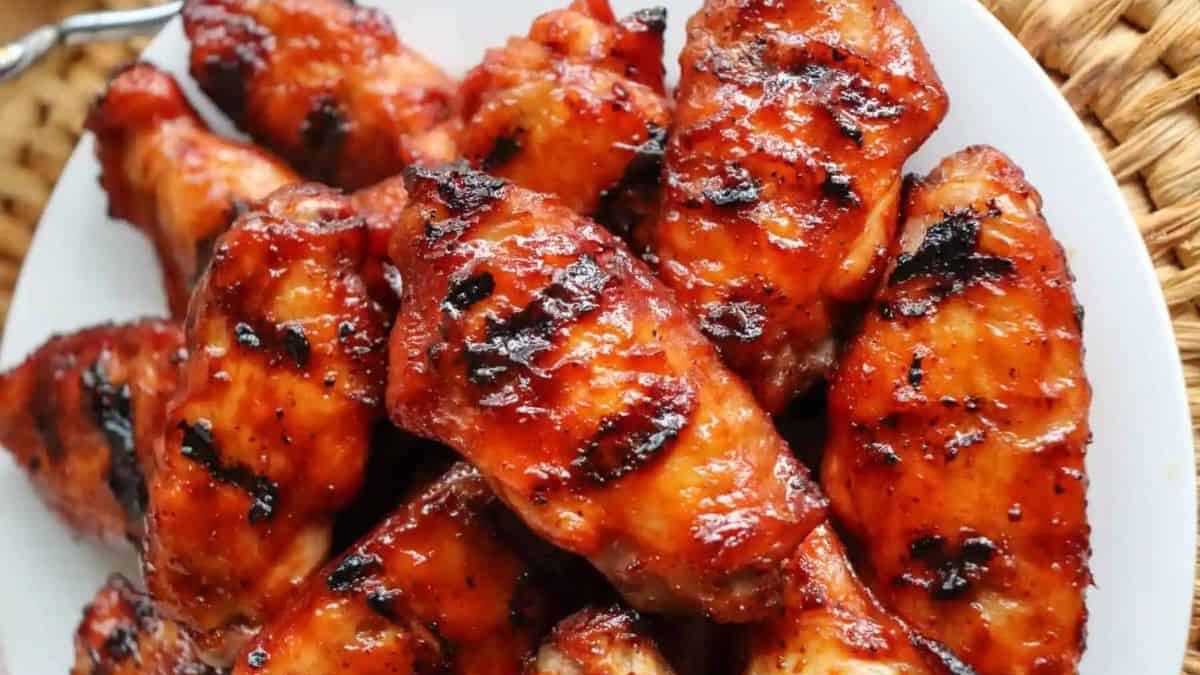 A plate of glazed, grilled chicken wings, crispy and golden brown, sits on a white dish. The chicken is coated in a shiny barbecue sauce, with charred grill marks visible. The background includes a woven surface partially visible.