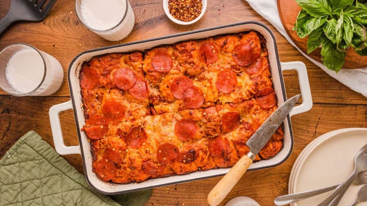 A freshly baked pizza cut into square slices in a casserole dish, surrounded by oven mitts, a spatula, glasses filled with milk, and plates with forks placed as decoration.