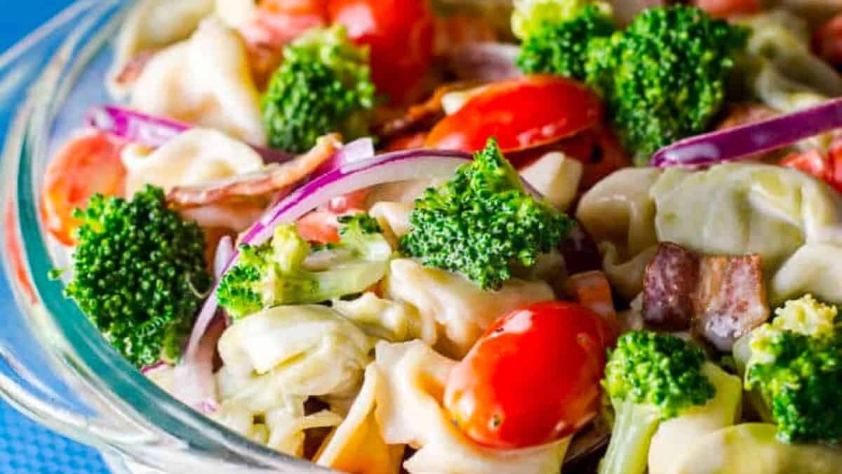 A vibrant pasta salad in a clear bowl features tortellini, broccoli, cherry tomatoes, red onion slices, and bacon. The colorful ingredients are mixed together, creating an appetizing and fresh appearance.