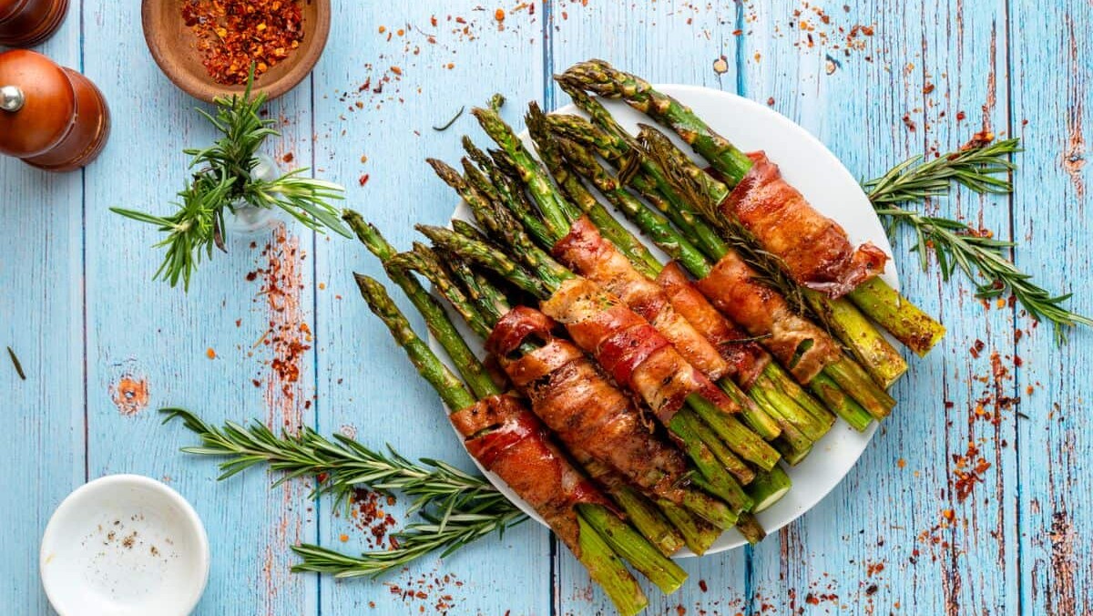 A plate of bacon-wrapped asparagus bundles is arranged on a rustic blue wooden table. Garnished with sprigs of rosemary and sprinkled with red pepper flakes, the dish is surrounded by small bowls and salt and pepper shakers.