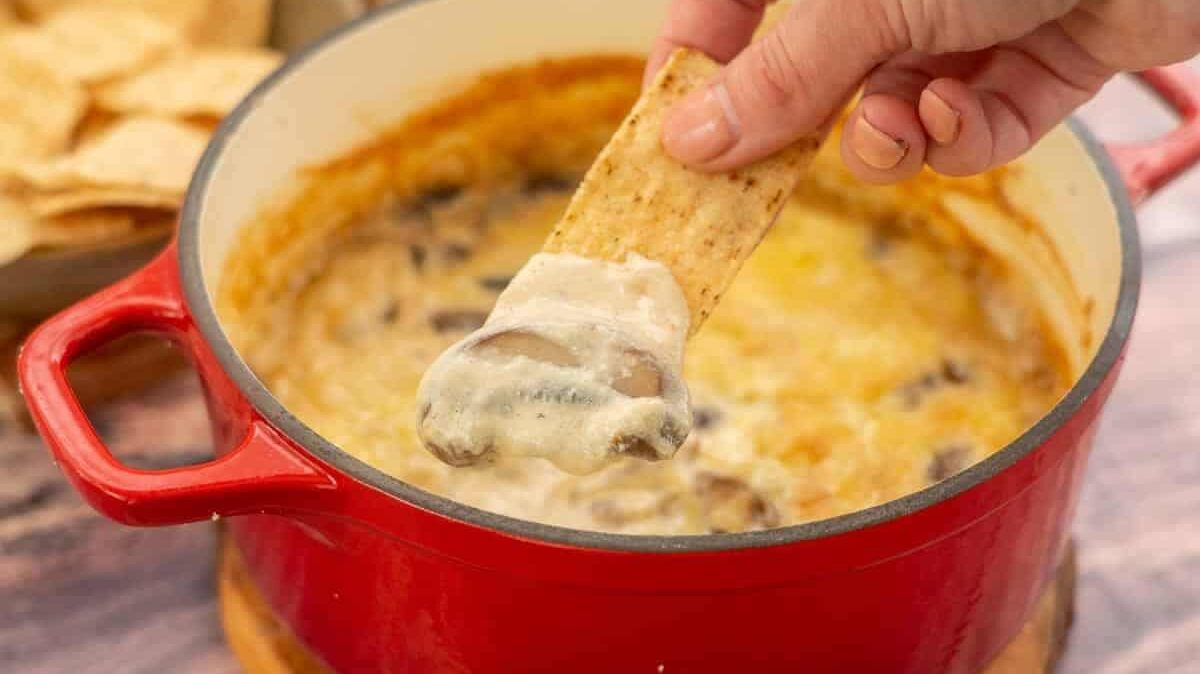 A hand dipping a tortilla chip into a red pot filled with creamy, cheese-covered mushroom dip. The pot is sitting on a wooden coaster, and some chips are visible in the background.