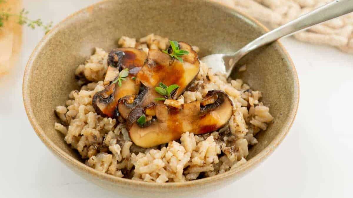 A bowl of mushroom risotto garnished with fresh thyme. A few sliced, browned mushrooms are on top, and a spoon is placed in the bowl. The setting is on a light background with a woven fabric napkin nearby.
